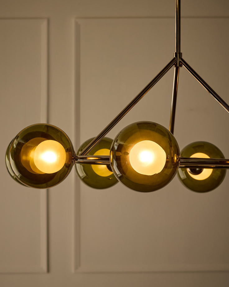 Riley 8-pendant light with olive-green glass globes and brass finish frame, two bulbs illuminated against neutral wall.