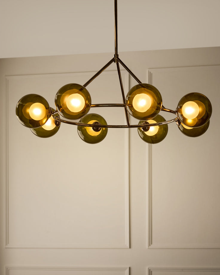 Riley 8-pendant light with olive-green glass spheres and dark metal frame, illuminated against white paneled ceiling.