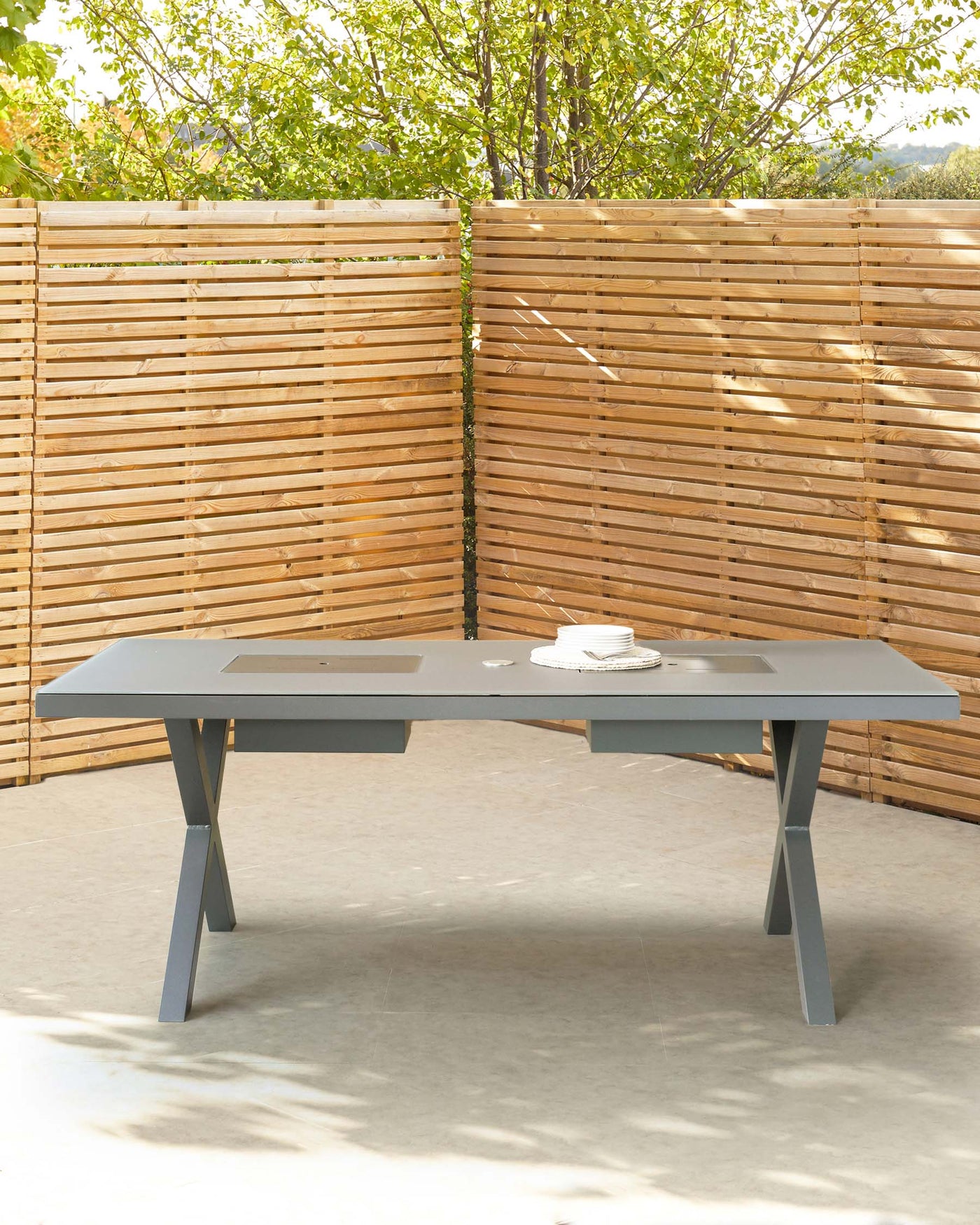 Modern outdoor rectangular dining table with a sleek grey finish, featuring angled legs for stability and a central cutout serving tray design. The table is set against a backdrop of wooden slatted fencing and lush greenery.