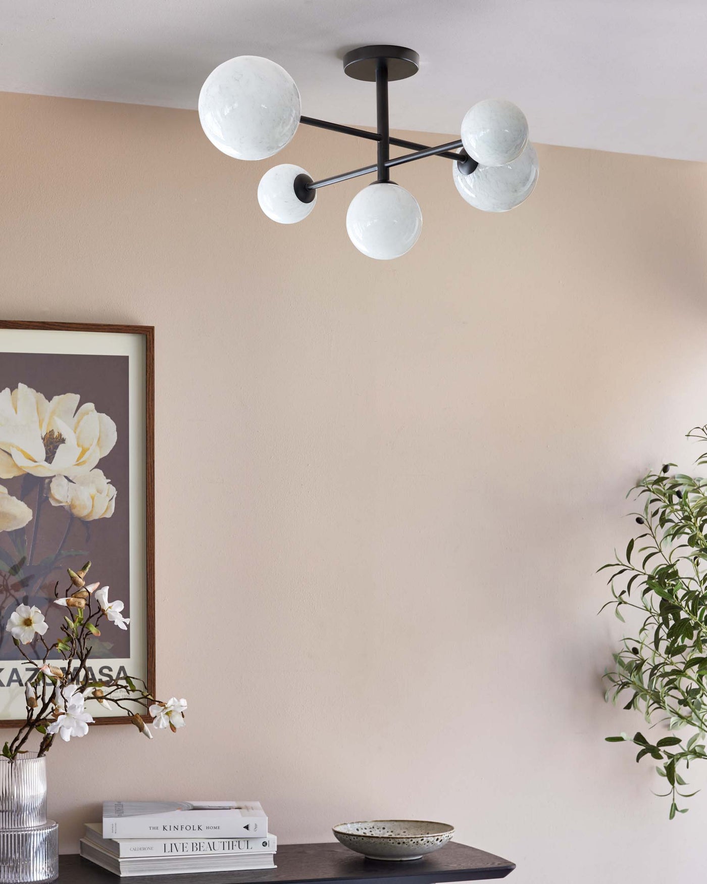 Modern black metal ceiling light fixture with multiple opaque white globes, set against a beige wall, above a dark wooden shelf displaying decorative items such as books, a flower vase, and a ceramic bowl.