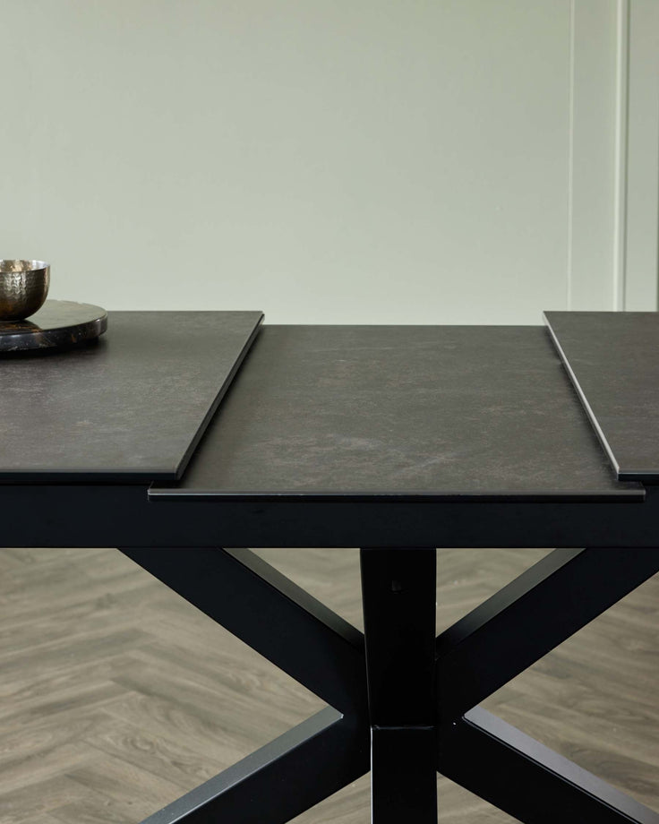 Stylish black dining table with a unique X-base and two extendable leaves, complemented by a decorative bowl and tray.