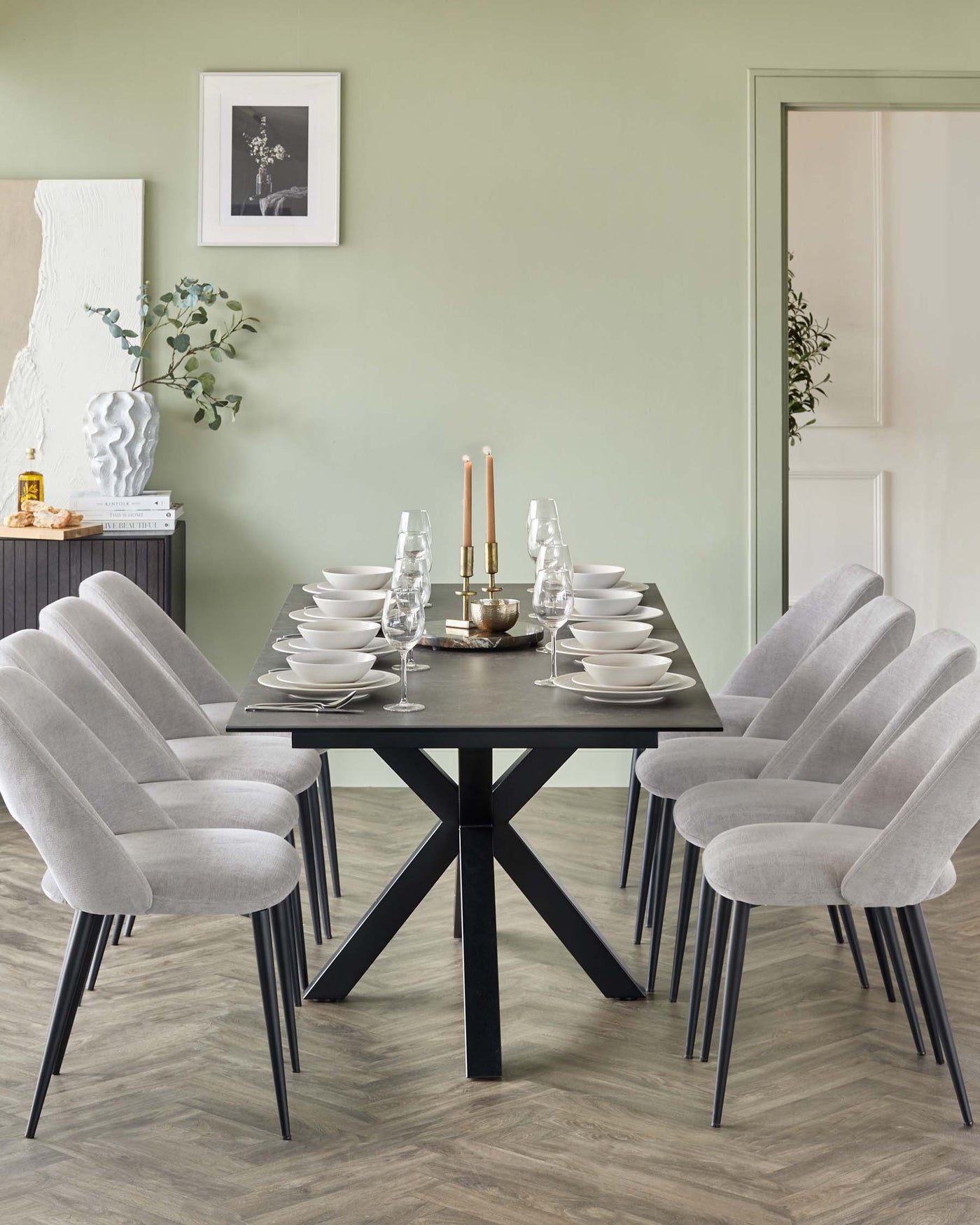 Modern dining set featuring a large rectangular black table with a X-shaped support base and six plush grey upholstered chairs with black legs positioned on a wooden floor. The table is set with white dishes, clear stemware, and a centrepiece with two tall candles.
