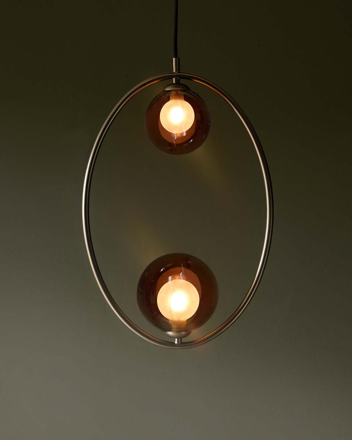 Oval pendant light with two amber glass globes suspended within a brushed metal frame. Modern lighting fixture.
