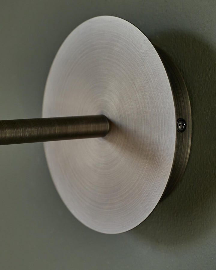 Close-up of a brushed metal wall sconce with a cylindrical arm, mounted on a green wall. Minimalist design.