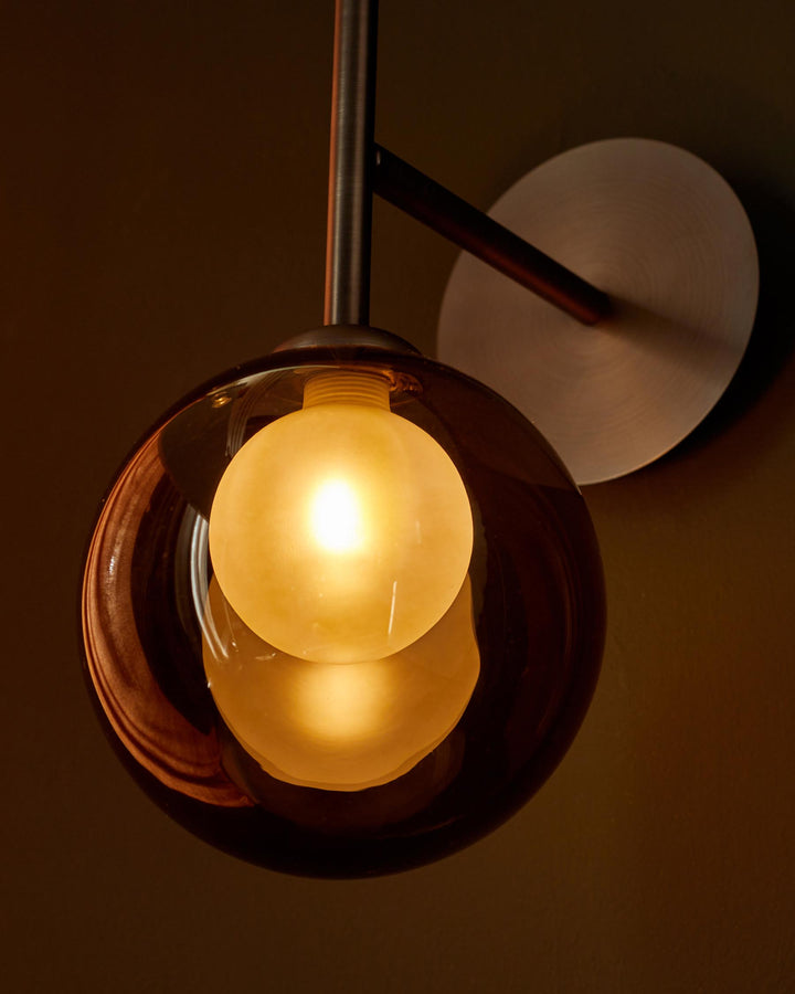 A stylish wall light fixture with a spherical glass shade and a brass-coloured backplate, casting a warm glow.