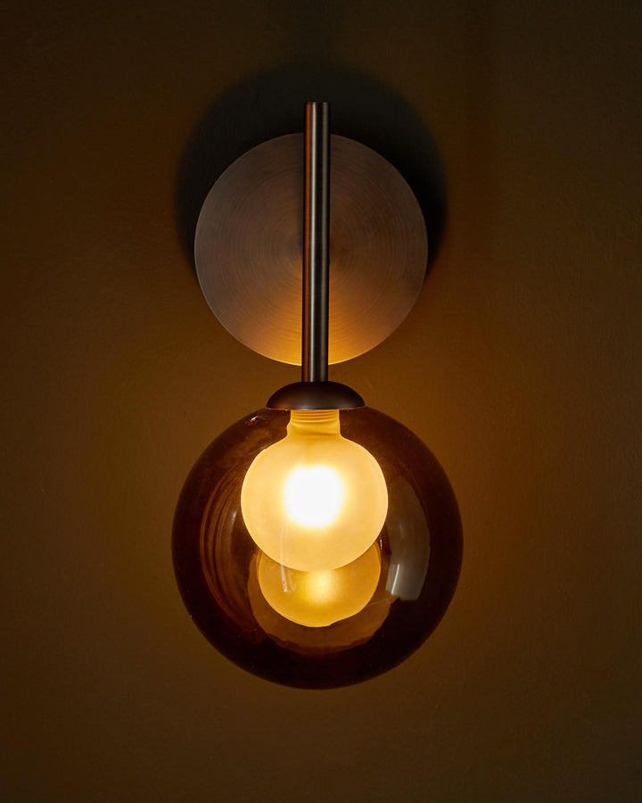 A modern wall light with a brass backplate and a spherical amber glass shade containing a glowing lightbulb.