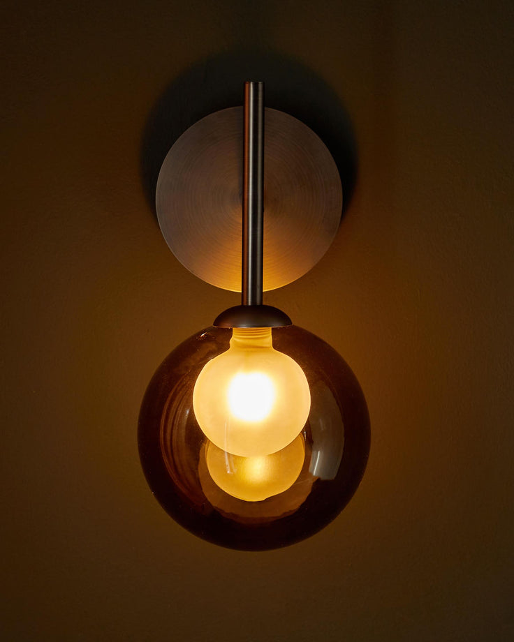 A modern wall light with a brass backplate and a spherical amber glass shade containing a glowing lightbulb.