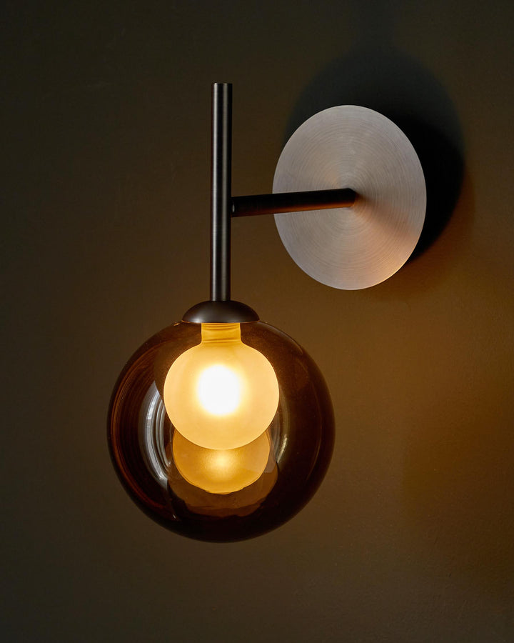 A stylish modern wall light featuring a spherical shade and brushed steel backplate, casting a warm glow against a dark wall.