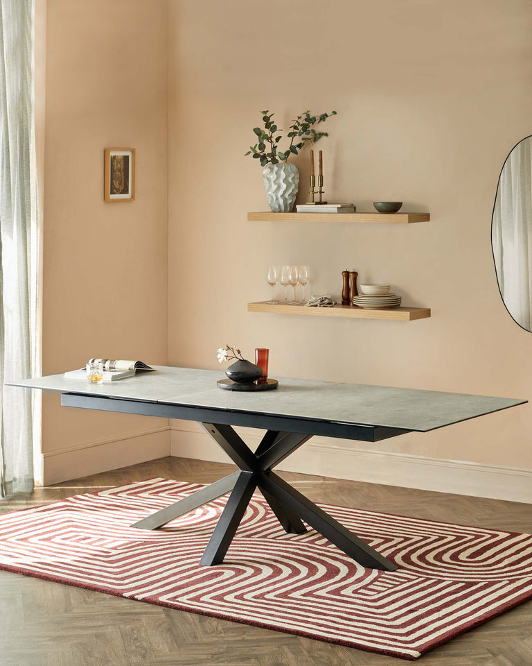 Contemporary rectangular dining table with a smooth grey surface and a bold black X-shaped base. Below, a vibrant red and cream patterned area rug adds a touch of colour to the wooden flooring.