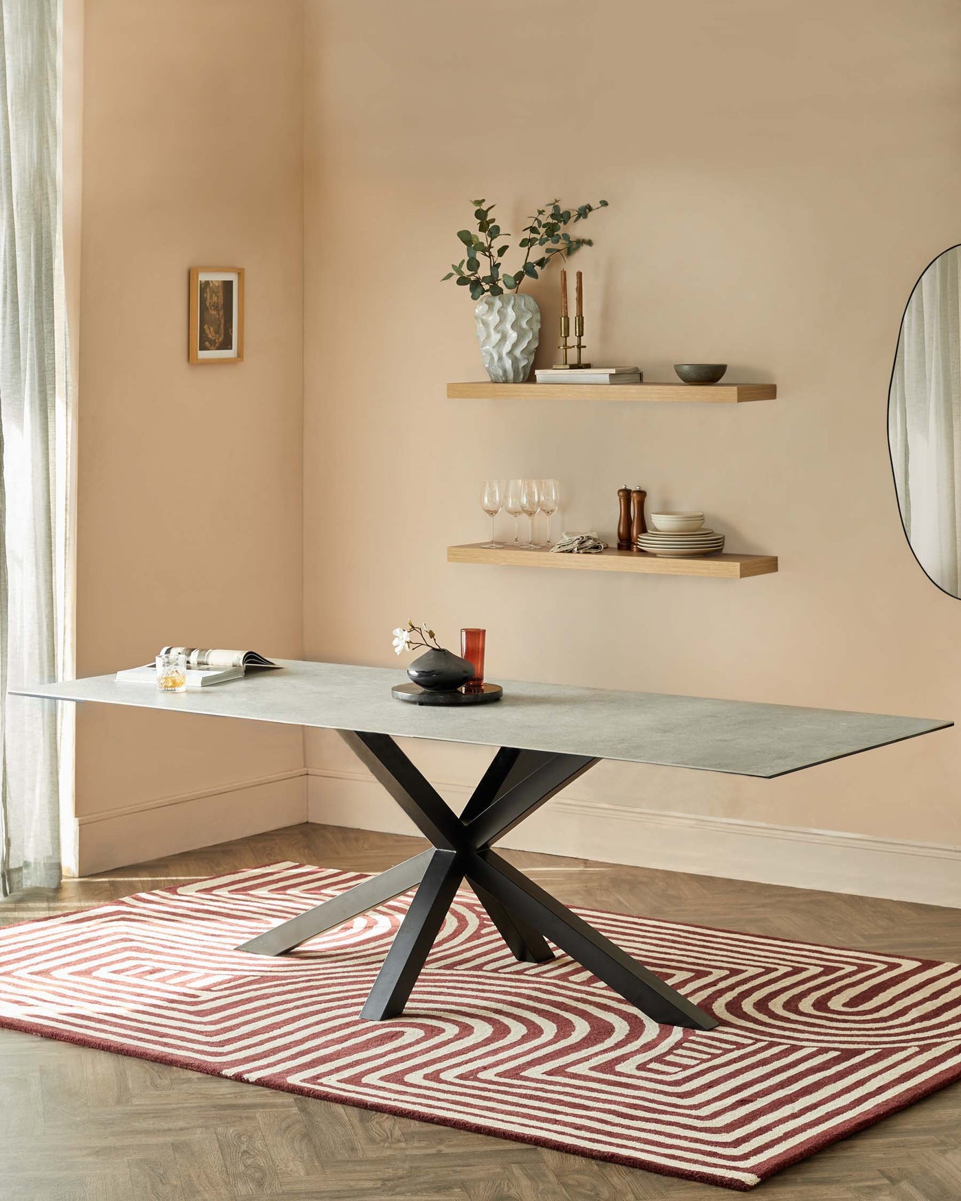 A modern rectangular dining table with a smooth grey surface and a geometric black base, accompanied by two floating wooden shelves on the wall above with decorative items, set against a warm peach-coloured wall background, and positioned on a red and white patterned area rug.