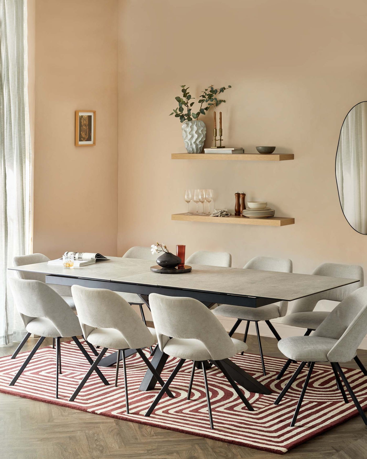A modern dining room setting featuring a sleek, rectangular table with a dark surface and black metal legs, surrounded by six mid-century-inspired upholstered dining chairs with light grey fabric and angled black metal legs. Above the table, two natural wood floating shelves display decorative items. The dining set is anchored on a geometric red and white patterned area rug.