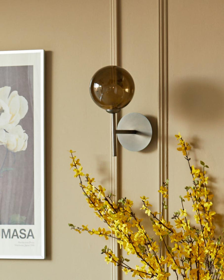 Stylish wall sconce with a brass finish and spherical smoked glass shade, next to a framed print and forsythia branches.