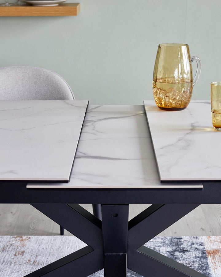 Modern dining table with a marbled white tabletop and a sturdy black cross-legged base, accompanied by a grey upholstered chair with a wooden shelf in the background. A textured amber glass pitcher and matching glass are placed atop the table. The setting is complemented by a patterned light and dark grey area rug beneath the table.