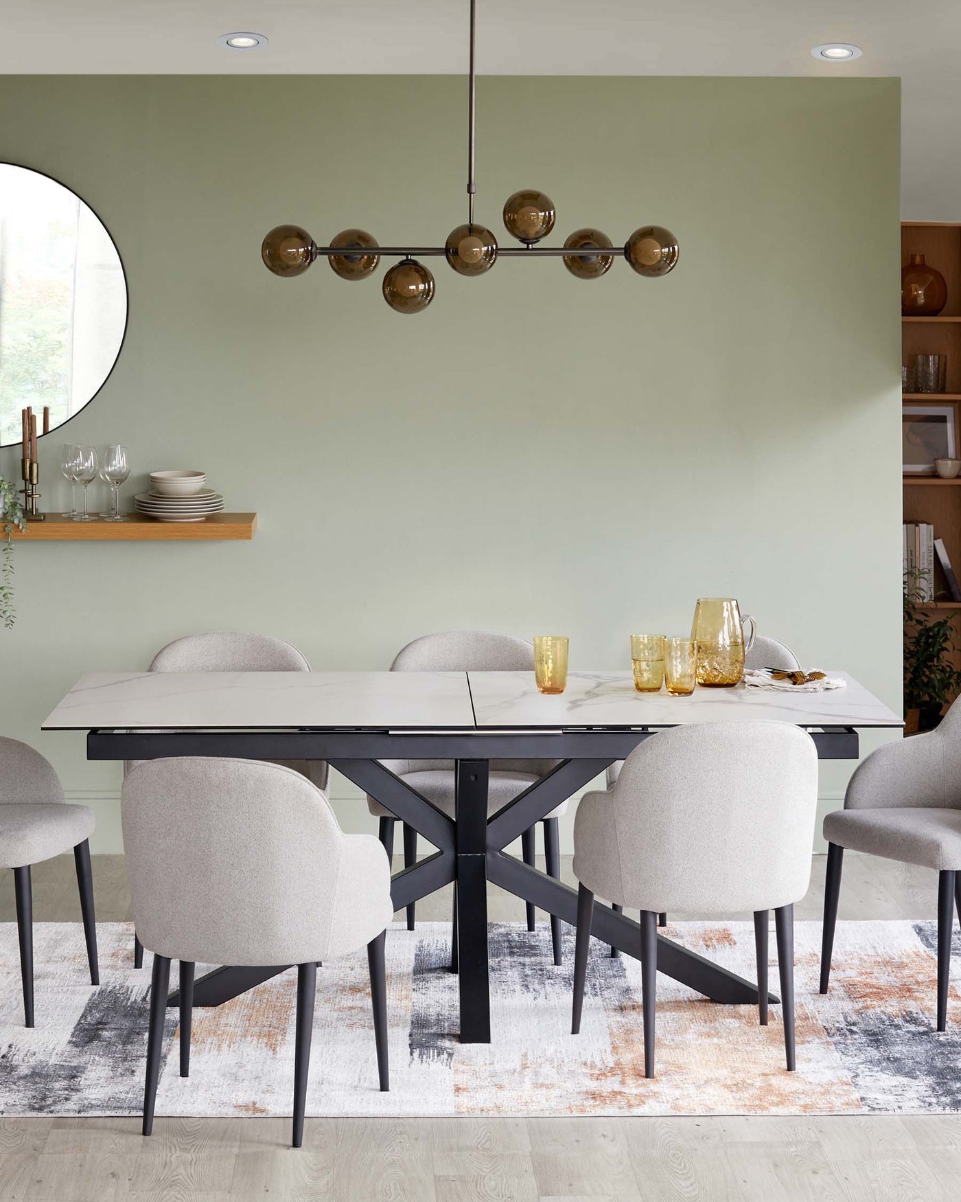 Modern dining room with an extendable black dining table featuring a white tabletop, surrounded by six grey upholstered chairs with black legs, over a multi-coloured area rug. A wooden shelf with dishes and a round mirror adorn the wall.