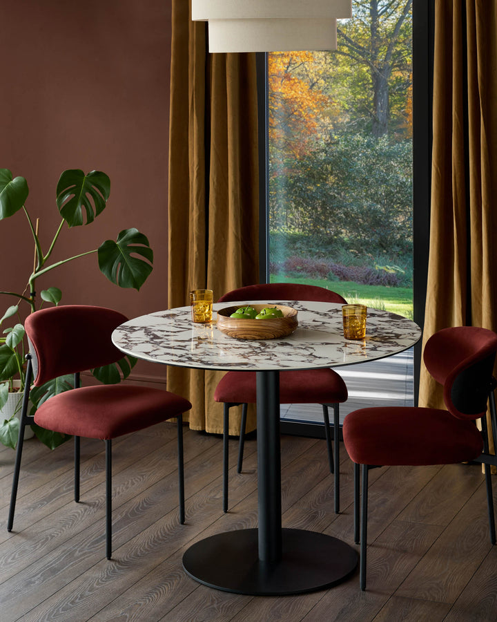 Dining area featuring a round marble table, four red chairs, a Monstera plant, and large windows overlooking greenery.