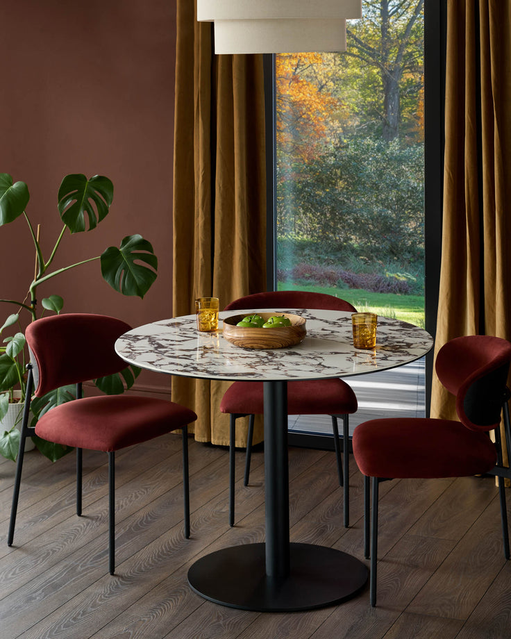 Dining area featuring a round marble table, four red chairs, a Monstera plant, and large windows overlooking greenery.