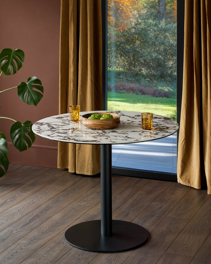 A marble-topped round table, with a black base, accessorised with drinks and a bowl of green apples.