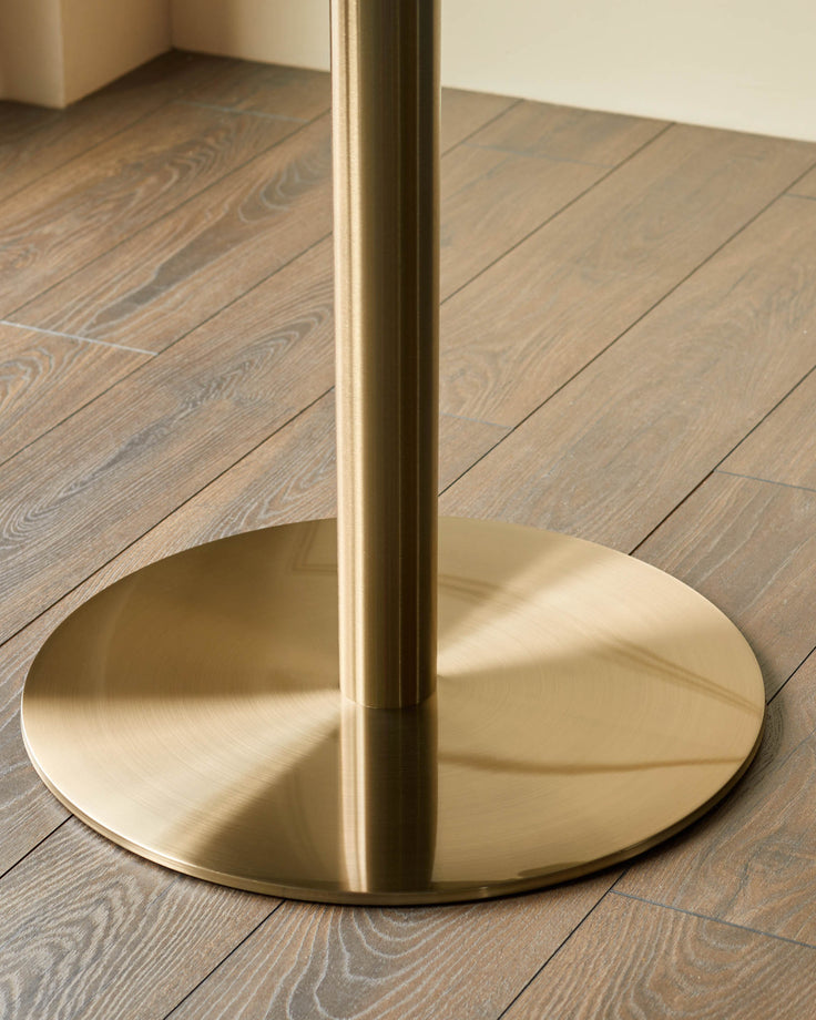 Close-up of a gold table base, showing a cylindrical column atop a circular, flat base, standing on a wooden floor.