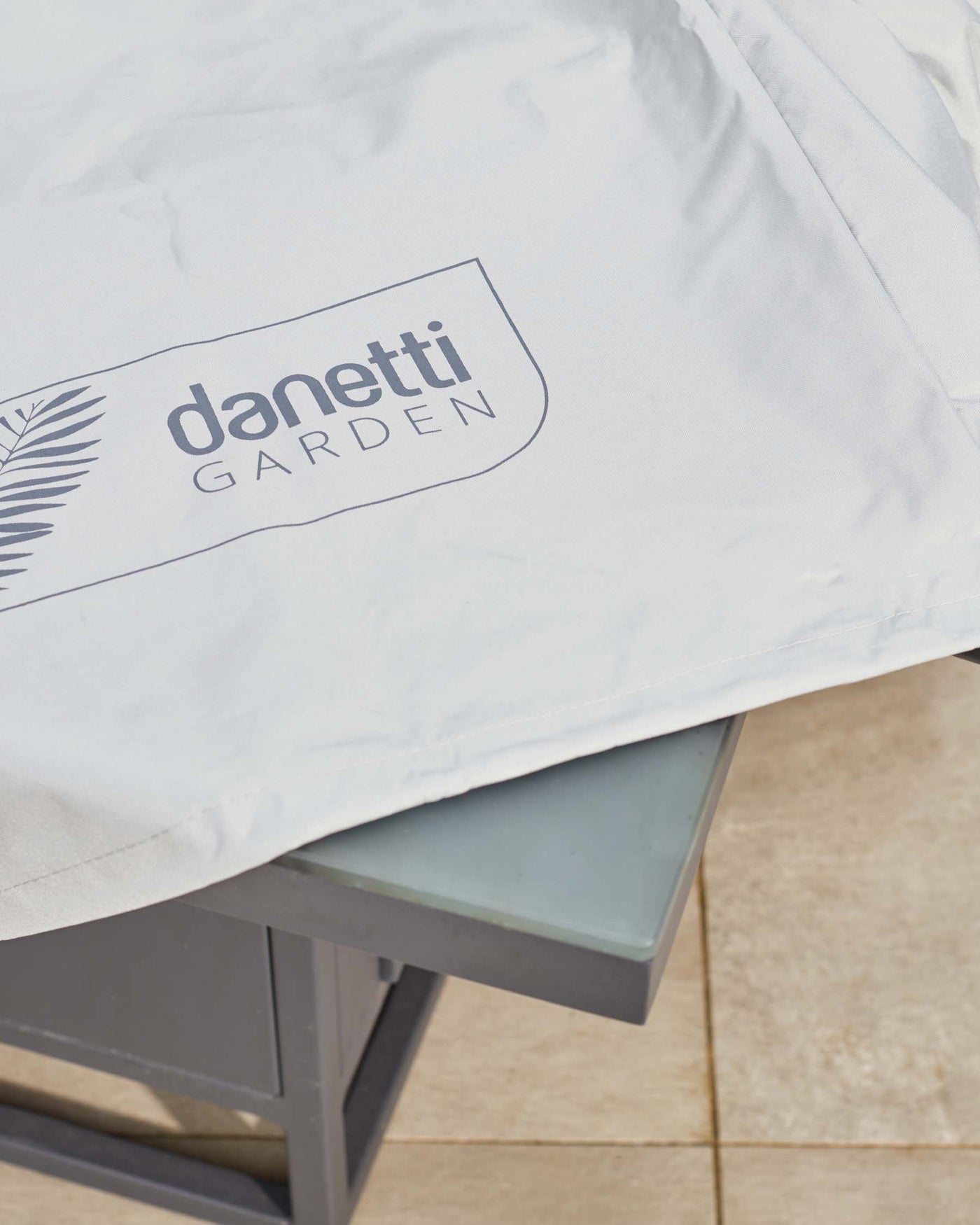 Close-up of a contemporary garden furniture set featuring a grey powder-coated aluminium table with a frosted glass tabletop and a white cushion with 'Danetti Garden' branding.