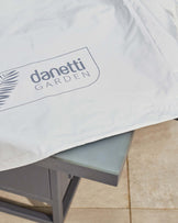 A grey outdoor table partially covered by a light-colored protective cover with the "danetti GARDEN" logo visible.