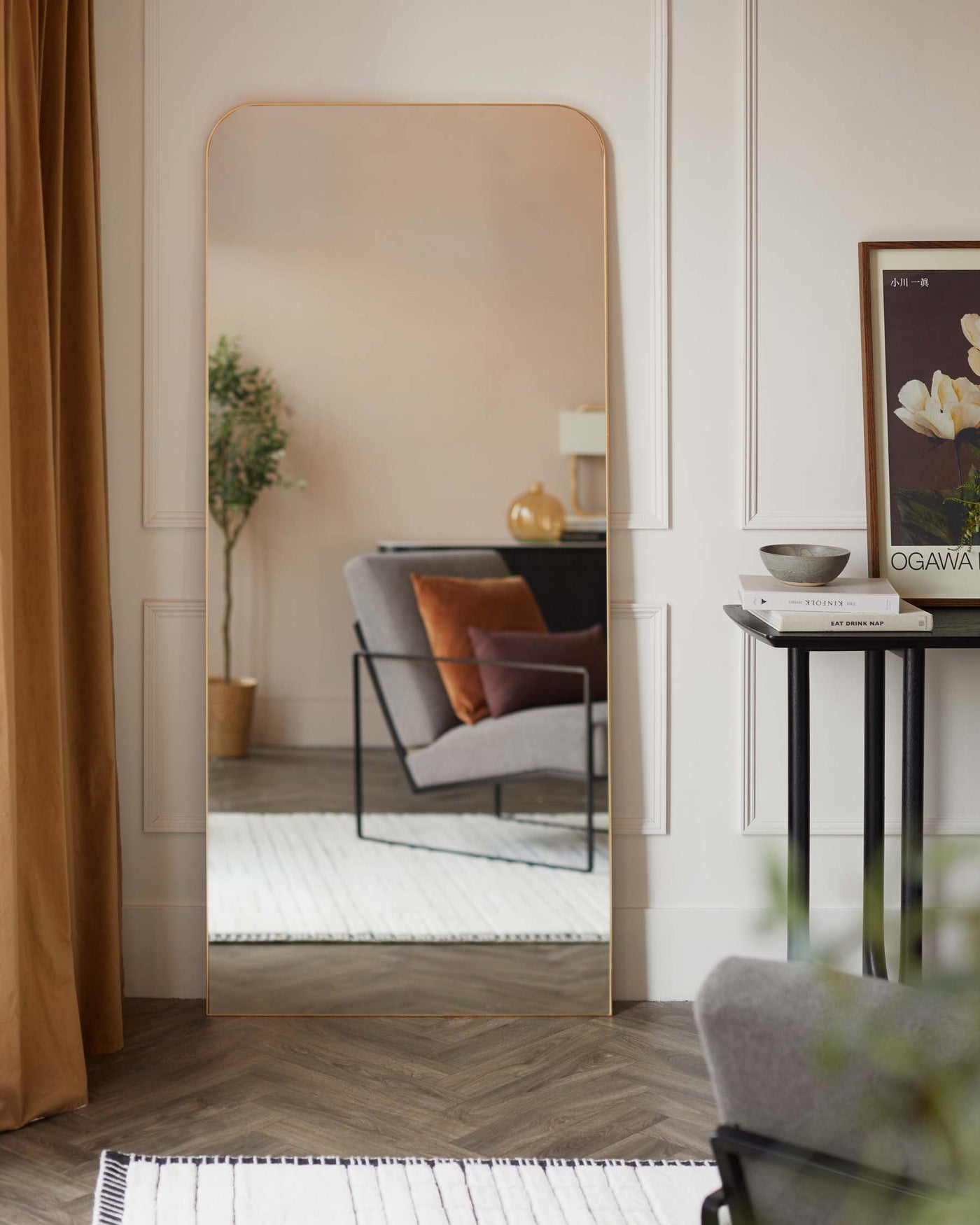 Elegant interior with a large, free-standing floor mirror with a minimalist rose gold frame. To the right, there is a sophisticated round side table with a black metal frame and dark tabletop, adorned with a small bowl and books. Reflected in the mirror is a stylish grey armchair with a black metal frame, complemented by an orange cushion. A framed picture, decorative vase, and potted plant add to the ambiance, creating a cosy and chic living space.