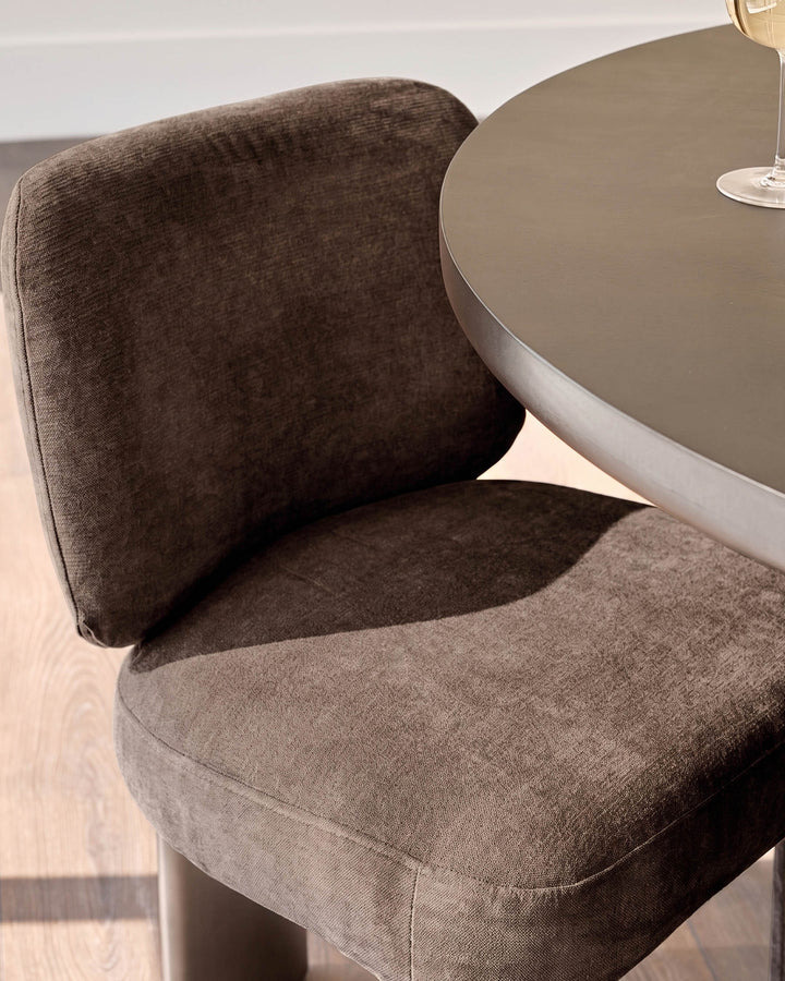 Rosalie dining chair in mocha fabric, shown at an angle next to a round taupe table, displaying curved backrest and seat cushioning.