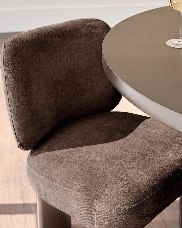 Rosalie dining chair in mocha fabric, shown at an angle next to a round taupe table, displaying curved backrest and seat cushioning.