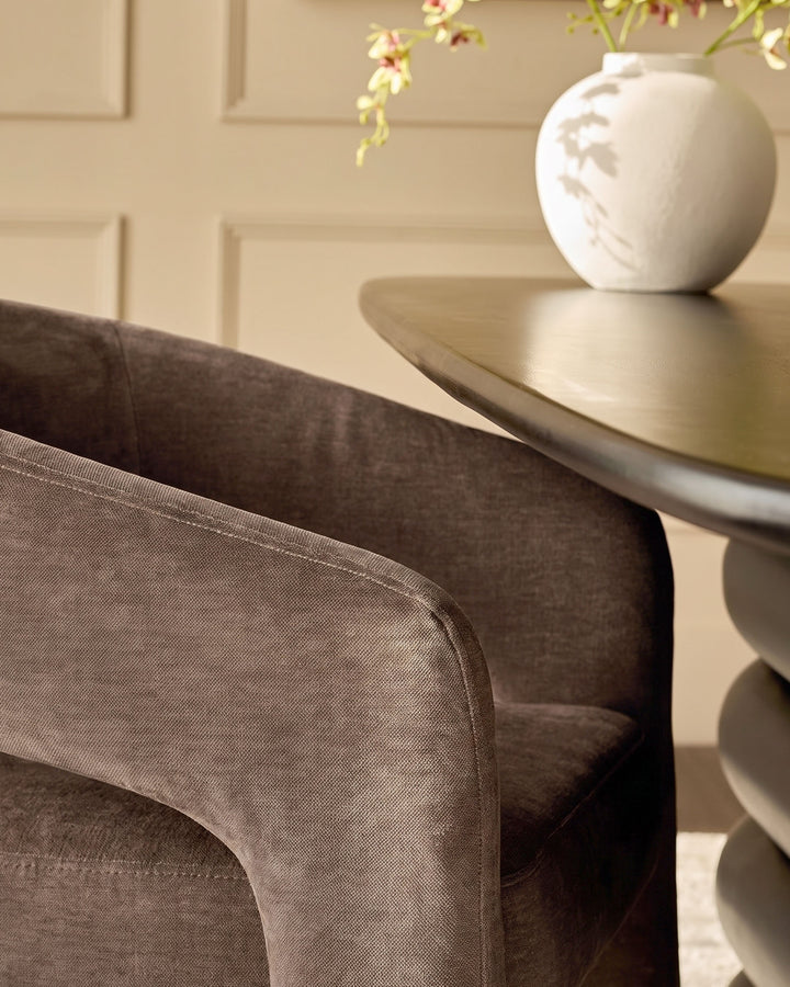 Rosalie dining armchair in mocha fabric, shown from a side angle next to a round white pedestal side table with flowers.