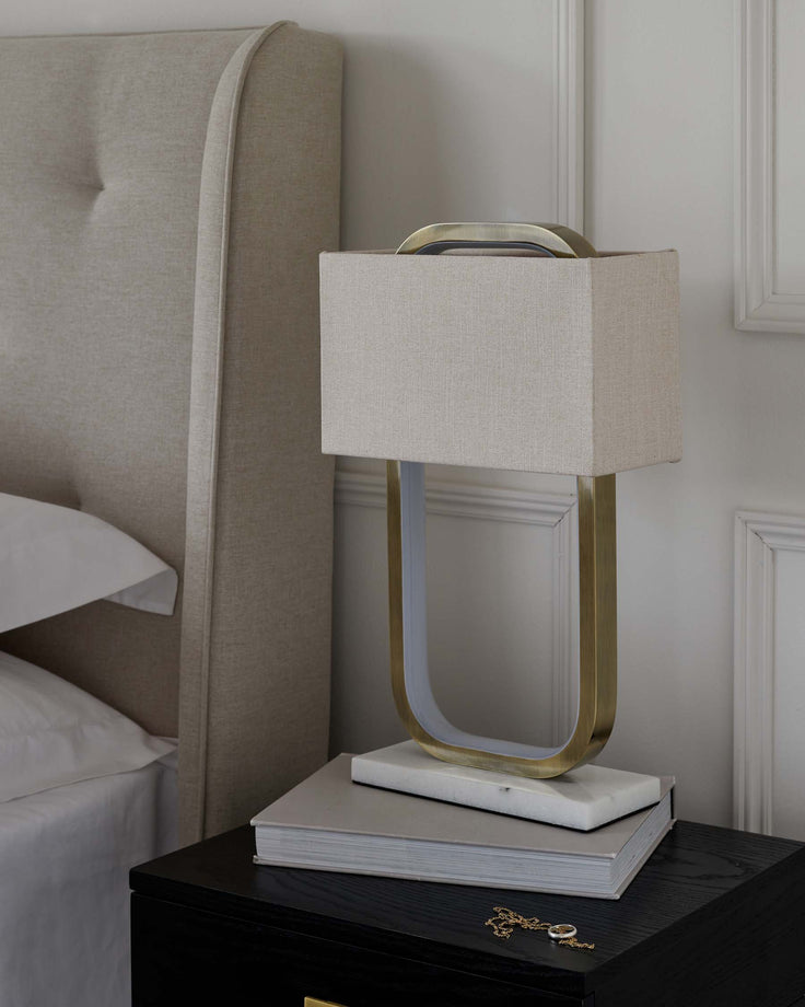 A modern black bedside table with a sleek finish and a brass handle is topped by a contemporary lamp with an elegant beige shade and a unique gold base featuring an open, geometric design. Adjacent to the lamp is a portion of an upholstered beige headboard with a curved silhouette and subtle tufting, suggesting a sophisticated, classic bedroom ensemble. On the table, beneath the lamp, lies a stack of hardcover books in neutral tones, adding a touch of intellectual charm to the scene.