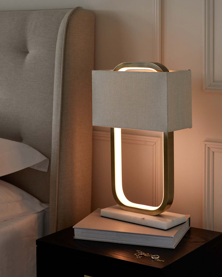 Elegant bedside setup featuring a modern table lamp with a distinctive U-shaped light source and a rectangular linen shade, placed atop a glossy black nightstand. Adjacent to the lamp is a pair of stacked white books and a delicate silver necklace, all next to a luxurious, upholstered headboard in a neutral beige tone.