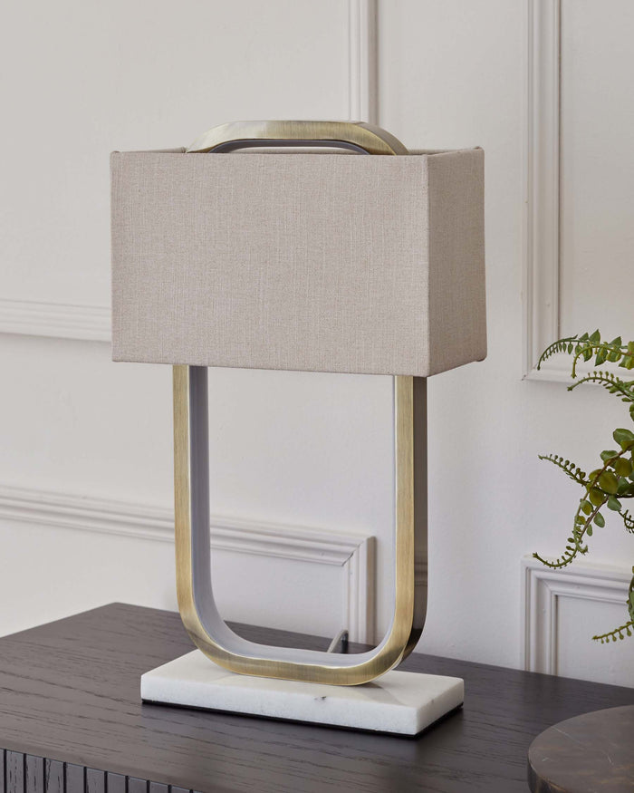 Modern table lamp with an open geometric brass base and a rectangular beige fabric shade, set on a white marble square base. Positioned on a dark wooden table against a white wall with decorative moulding.