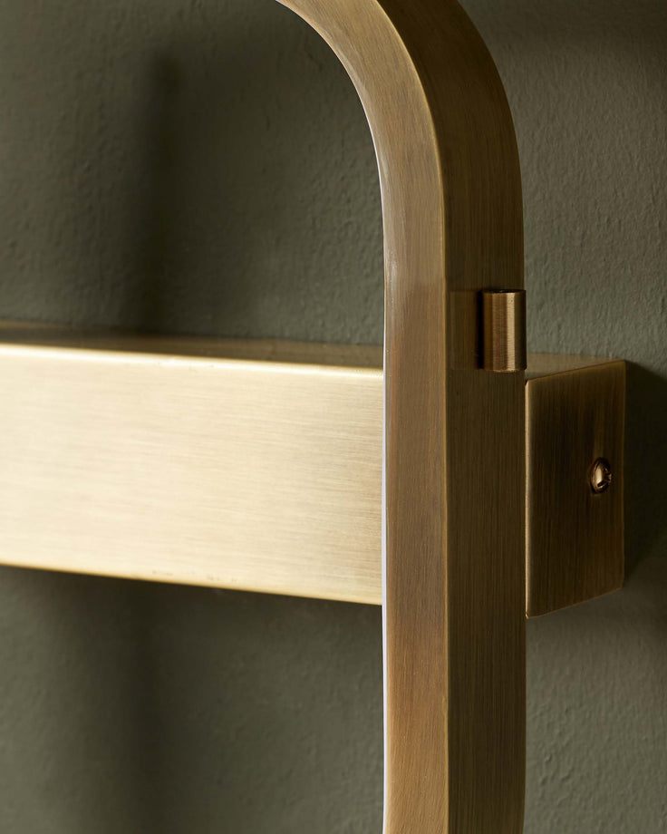 Close-up of a brass towel rail mounted on a textured, dark green wall. The rail is made of bent and rectangular metal pieces.