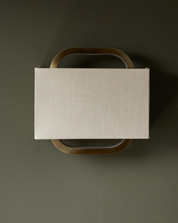 A rectangular, fabric lampshade affixed to a brass frame, mounted on a textured, olive-green wall.