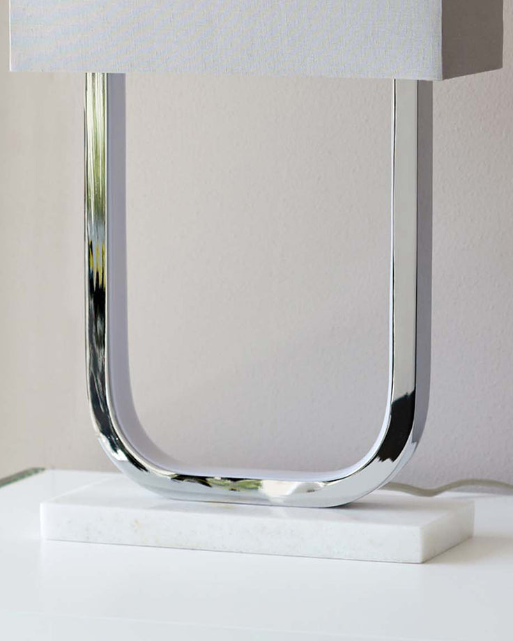 Sanza chrome table lamp with U-shaped chrome base and white marble base, displayed against a neutral background.