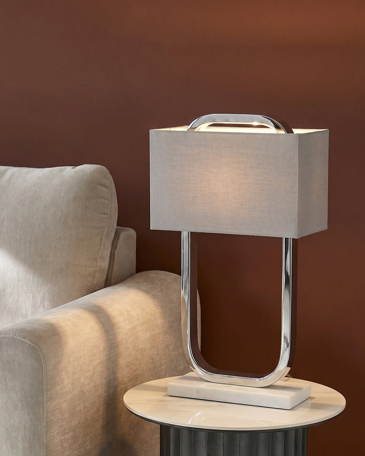 Sanza chrome table lamp with white linen shade, curved metal base, displayed on round marble side table next to beige armchair.