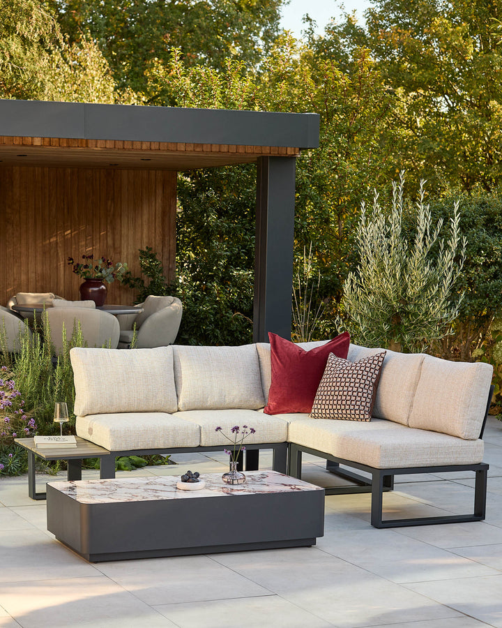 Savannah large corner bench set with black base, natural cushions, red and patterned pillows, and marble-top coffee table in garden setting.