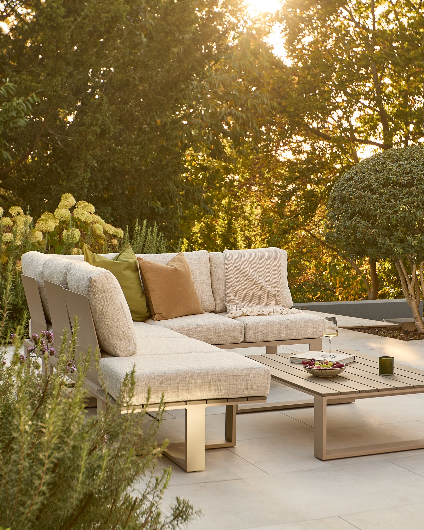 Savannah garden large corner sectional in natural with bench coffee table set in outdoor patio setting with trees.