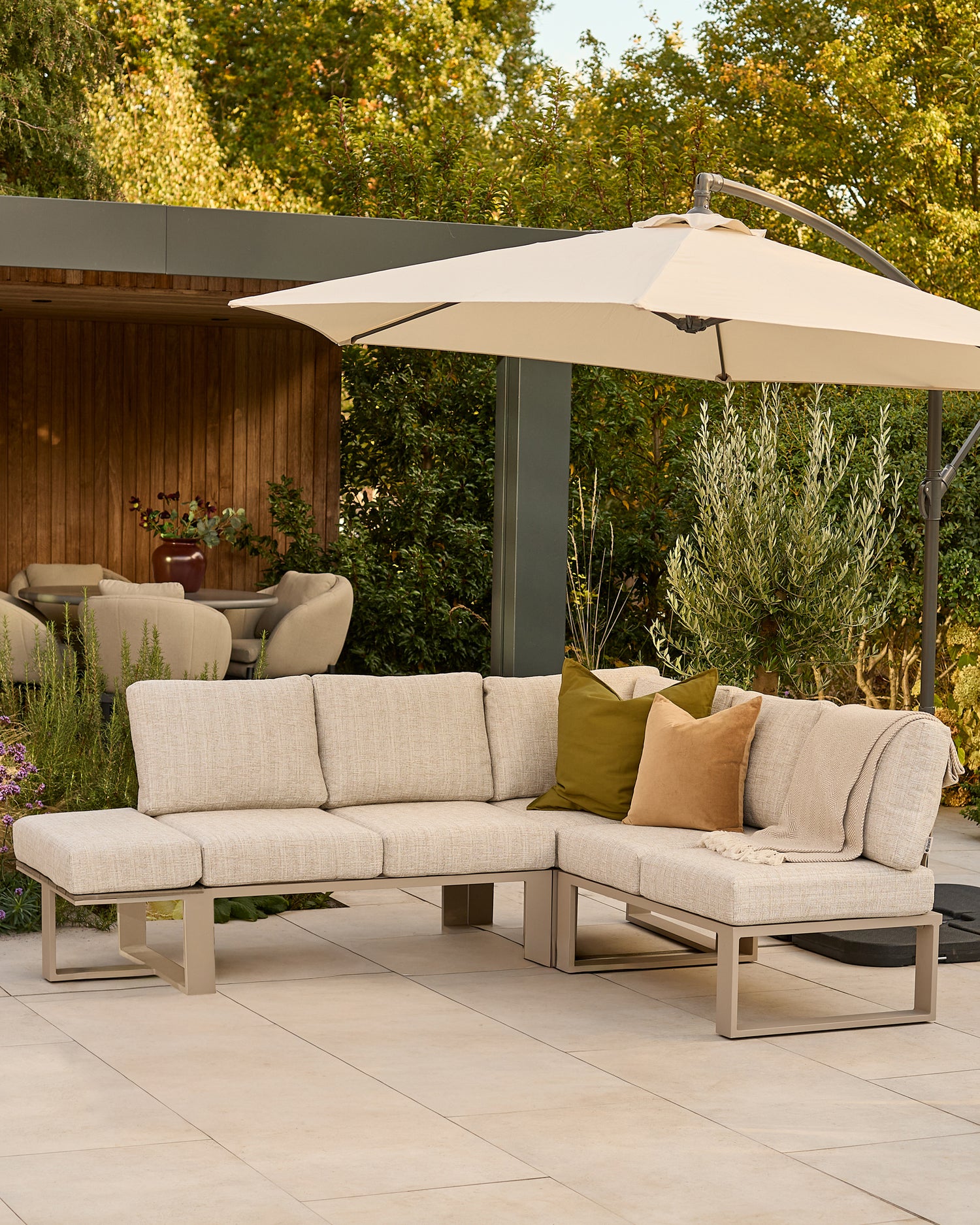 Savannah large corner garden bench with natural base, beige cushions, styled under cream umbrella in outdoor patio setting.
