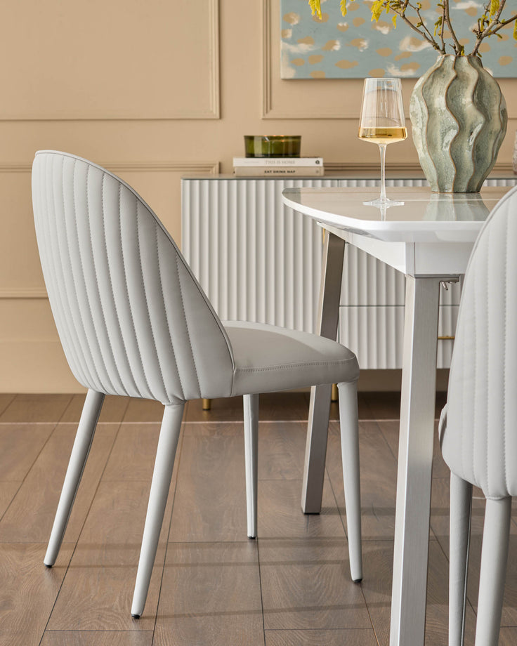 Saylor light grey faux-leather dining chair with vertical quilted backrest and white metal legs, shown at white table.