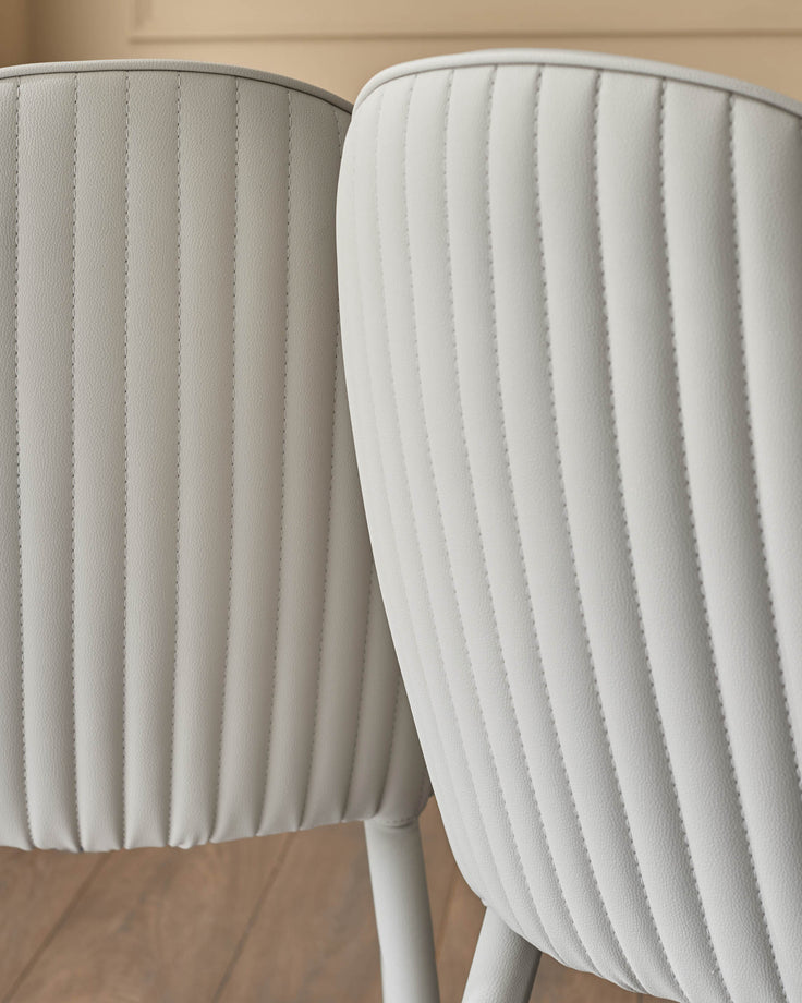 Two light grey faux-leather dining chairs with vertical stitched paneling and curved backrests, shown from a close-up side angle.