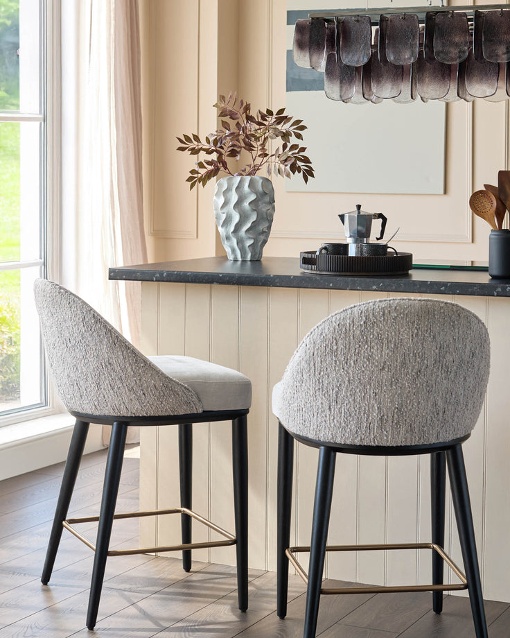 Two stylish bar stools with textured grey fabric, dark legs and brass footrests sit at a kitchen counter.