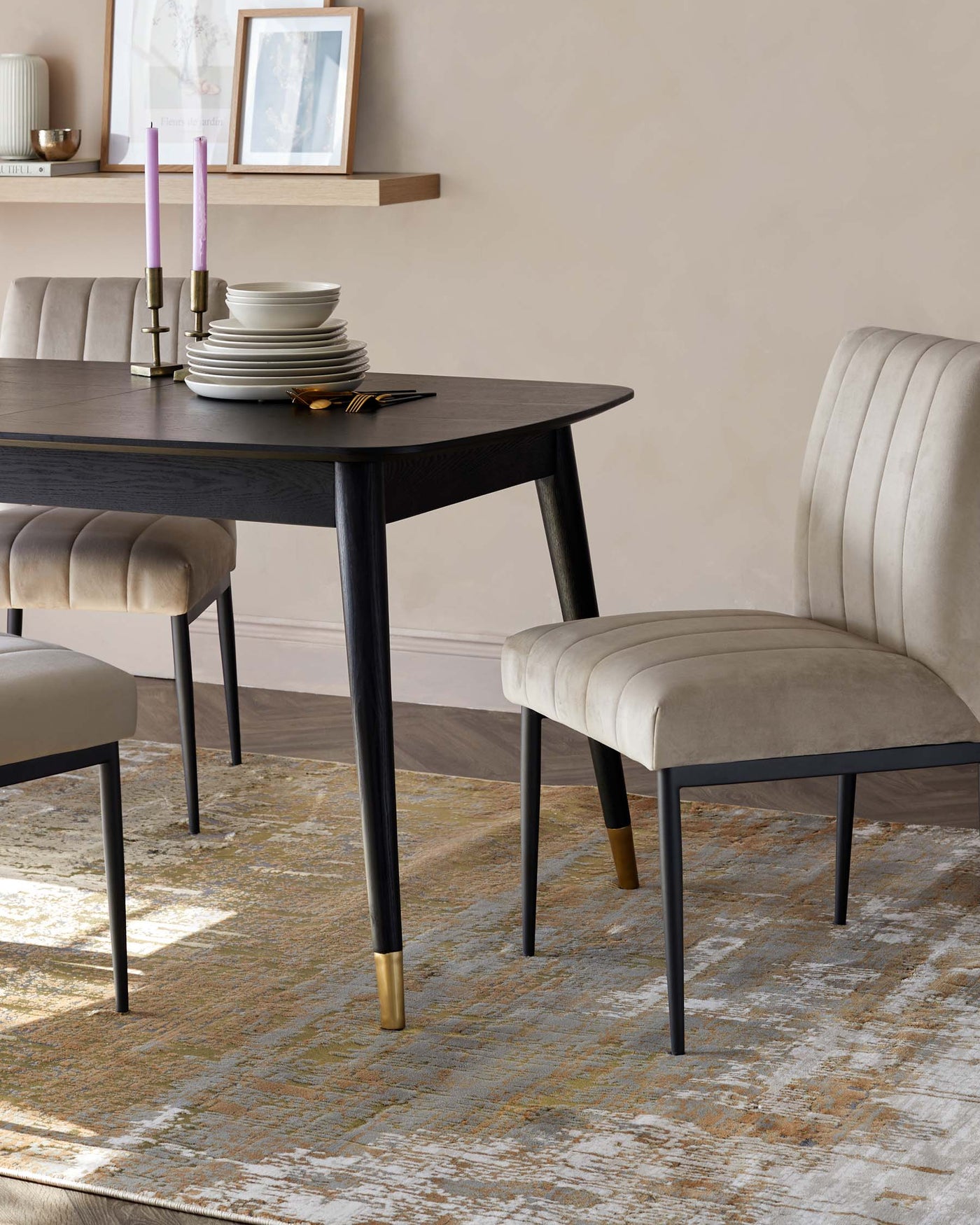 Elegant dining room setup featuring a sleek, black rectangular table with tapered legs finished with gold-coloured tips. Accompanied by three plush, velvet, beige dining chairs with subtly curved backs and black, slender legs, matching the table. The ensemble is positioned on top of a distressed, multi-coloured area rug, blending warm hues with a touch of cool tones.