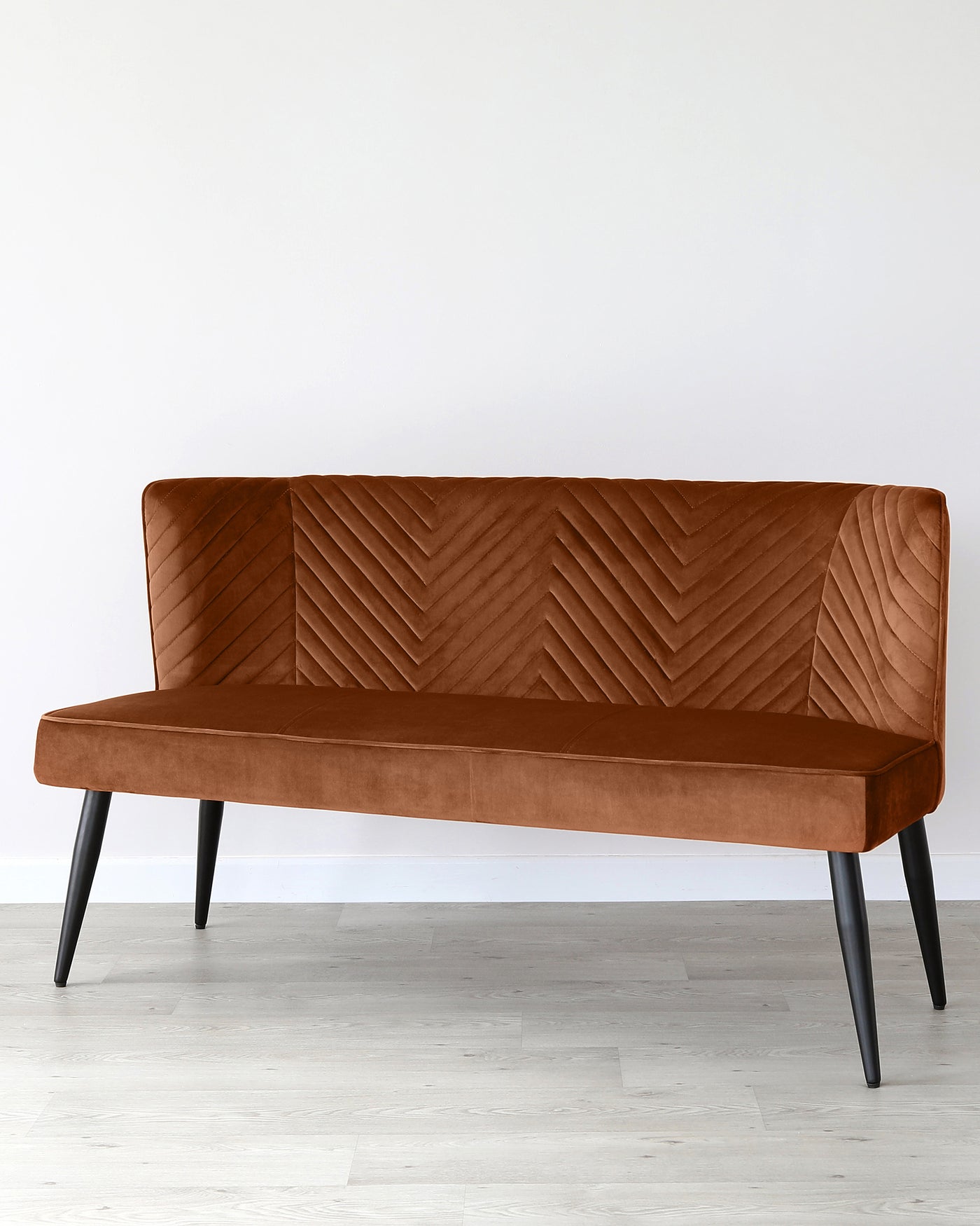Modern mid-century style bench with a high backrest and chevron pattern upholstery in a warm caramel tone, supported by sleek tapered black legs, set against a clean white wall on a light wood floor.