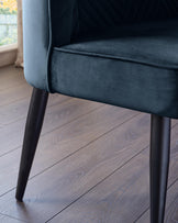 Close-up of an upholstered chair in teal velvet with dark, slender legs, sitting on a wooden floor near a window.