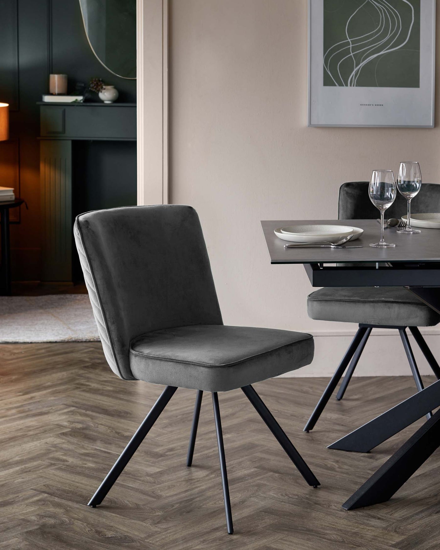 Modern dining room with a grey upholstered chair featuring a smooth, curved backrest and cushioned seat on angled black metal legs. In the background, there's a black contemporary dining table with a tempered glass top and unique geometric black metal legs, partially set with white plates and clear wine glasses.