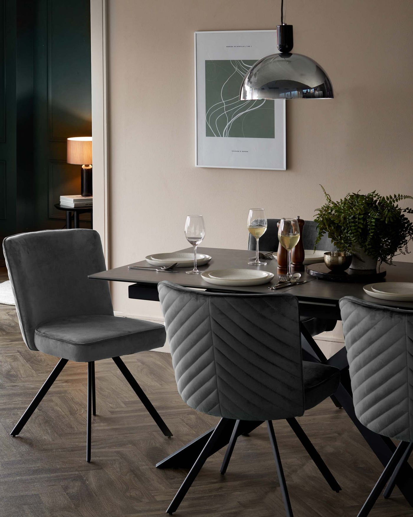 Contemporary dining furniture set featuring a sleek round table with a dark tabletop and angular black legs, accompanied by four plush, grey, velvet upholstered chairs with chevron-stitch detailing and splayed metal legs. A modern pendant light with a chrome finish hangs above the table, accentuating the sophisticated, minimalist decor of the dining space.