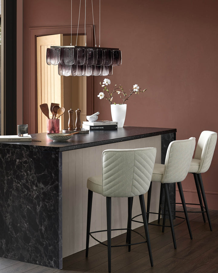A kitchen island with 3 upholstered bar stools. Overhead is a cascading, smoky grey pendant light fixture.