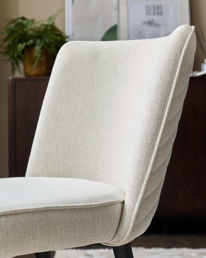 A stylish cream upholstered chair with a ribbed back, paired with a dark wooden cabinet and a plant.