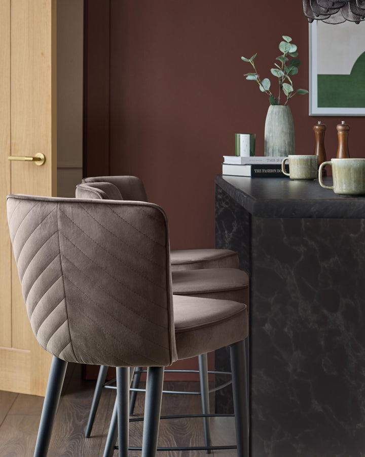 Mocha velvet bar stool with quilted backrest and dark metal legs, positioned at black marble counter in modern interior.