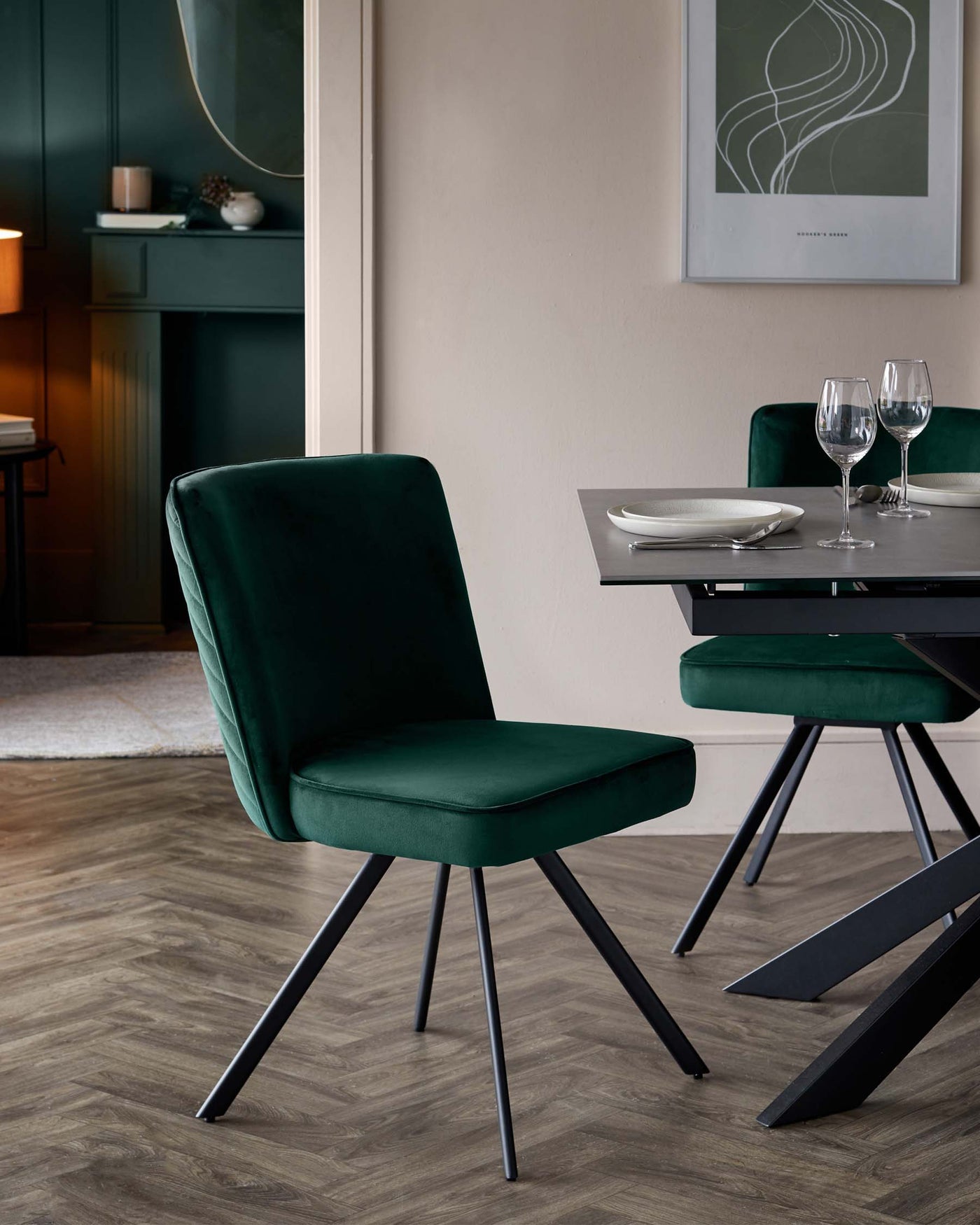 Plush green velvet dining chair with a sleek, vertical tufted backrest and cushioned seat, resting on angled black metal legs. Contemporary dark-toned wooden dining table with geometric black metal base, partially visible. Two clear wine glasses sit on the table, suggesting a refined dining setup.